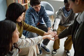 Group of diverse teenagers sitting in counseling session joining hands in center showing teamwork and support, multiethnic boys and girls participating in group therapy activity