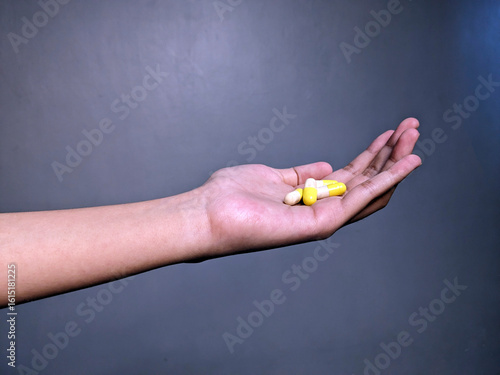 A person's open palm holds two yellow and white capsules, presenting medication or supplements against a neutral gray background.