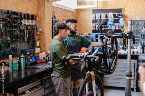 Two mechanics repairing bicycle wheel in workshop using laptop