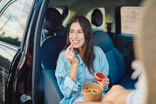 Smiling young woman enjoying french fries with ketchup, sitting in passenger seat with open door