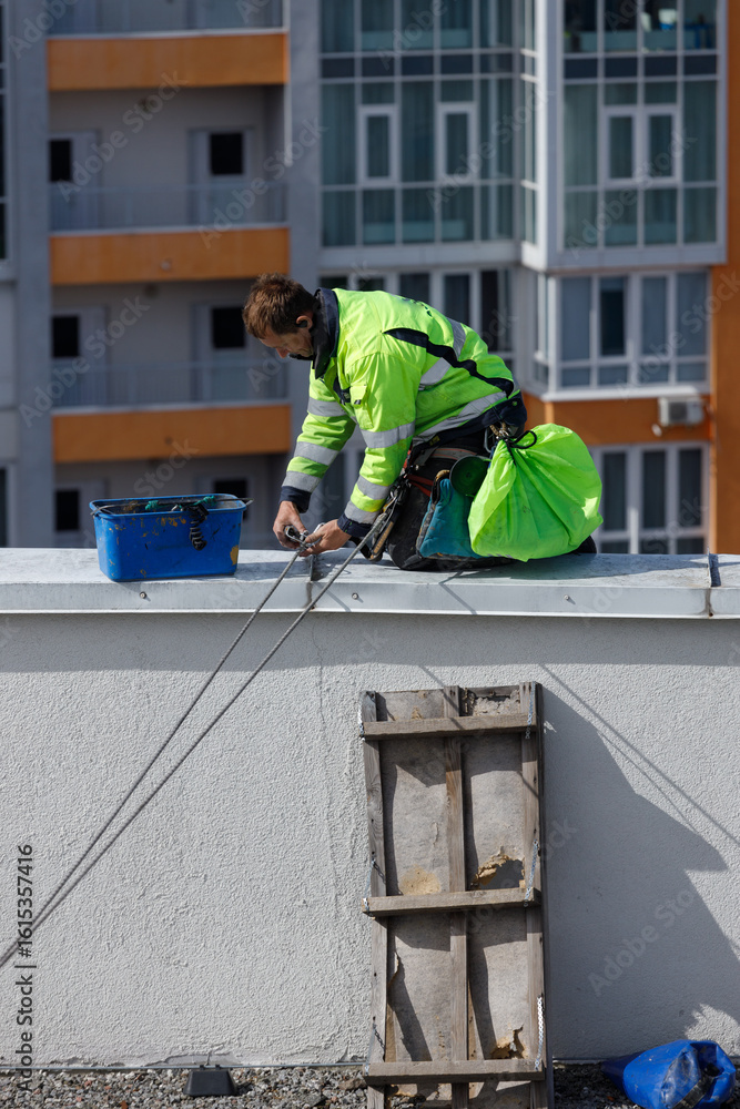 Fototapeta premium Construction Worker Performing Maintenance on Rooftop While Wearing Safety Gear in Urban Setting with Modern Apartments in Background Under Clear Blue Sky
