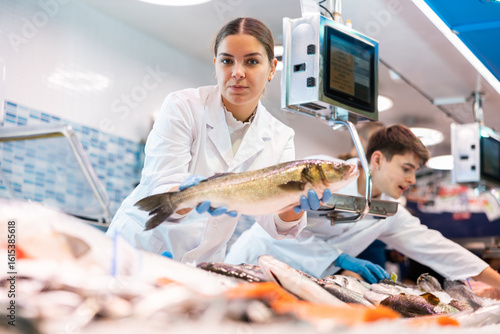 Valokuva Female seller in apron standing near counter offering fresh fish hake to the buy