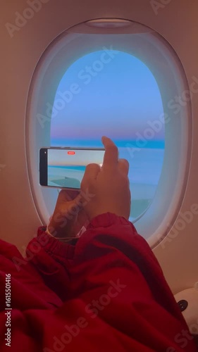 Tourist woman hand used smartphone taking footage vdo through airplane window of Pastel pink cloud in morning sunrise blue sky on aircraft wing on holiday vacation travel trip, 4k vertical aerial view