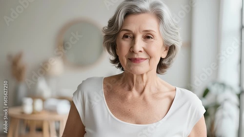 Beautiful gorgeous 50s mid aged mature woman looking at camera An older woman with gray hair smiles warmly in a softly lit, cozy, modern living space.