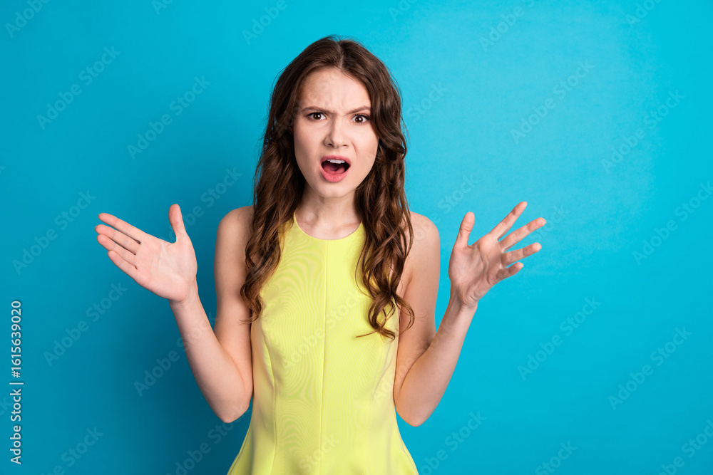 Fototapeta premium Young charming woman in stylish yellow dress on blue background, expressing surprise with raised hands, promoting lifestyle