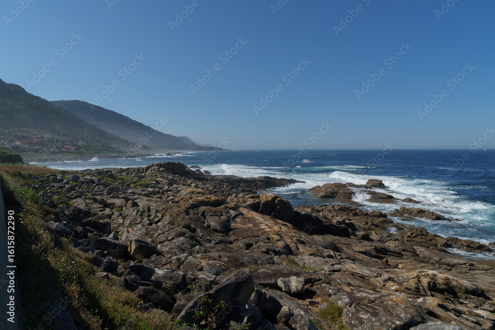 Obraz premium Scenic view of powerful ocean waves crashing against a rocky coastline, with majestic mountains rising in the distance under a clear blue sky
