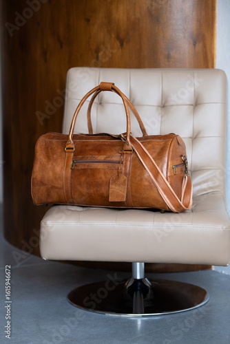 A vintage-style brown leather duffel bag rests on a cream-colored tufted chair against a blurred wooden and grey background