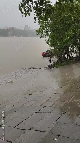 Atmospheric scene of a monsoon rainstorm over a wide, murky river, with a small boat moored by a wet stone ghat and lush green trees 4k video