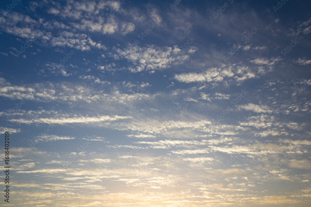 Fototapeta premium Golden sunrise with delicate scattered clouds in a serene blue sky, capturing the peaceful beauty of early morning light and atmospheric texture.