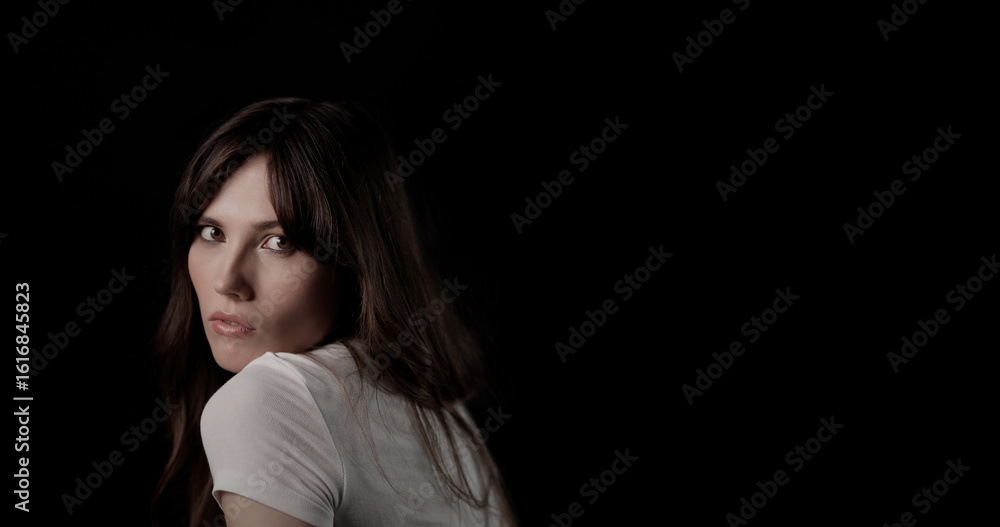 Obraz premium Beautiful young woman with long brown hair wearing white t shirt looking over her shoulder on black background