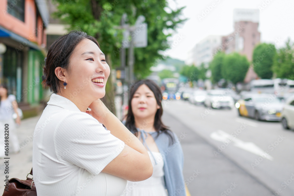 Obraz premium On a bustling boulevard, two Korean women in their twenties in casual clothes stand at a bus stop, chatting quietly as they wait for the bus under soft spring light. Seoul, Jongno District, spring.