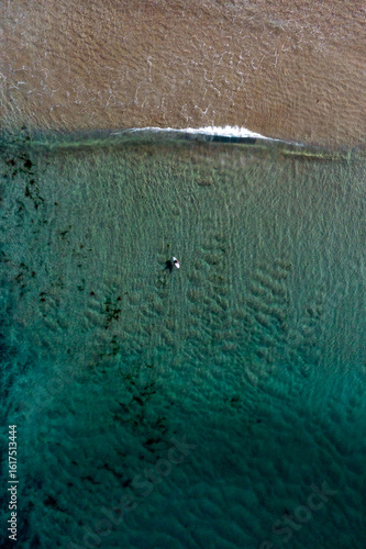 Idyllic, crystal clear, shallow turquoise ocean water lining a sandy shore with abstract sea foam textures from a breaking wave. Captured from high above the beach.