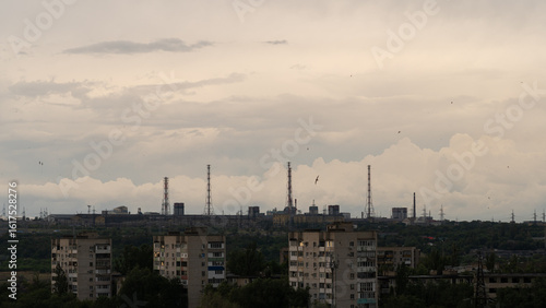Wide view of aging apartment blocks facing distant industrial complex with tall smokestacks and power lines, silhouetted against layered storm clouds at dusk