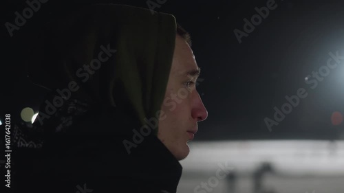 Side view of unhappy boy in hoodie standing outside during night with bright light glowing in distance, capturing mood of isolation and introspection in cold evening atmosphere