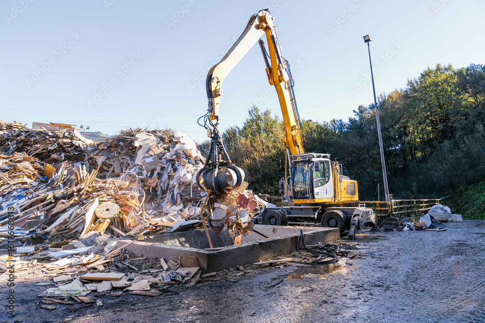 Fototapeta premium Excavator is working on a recycling site, picking up construction and demolition waste with its grapple for further processing