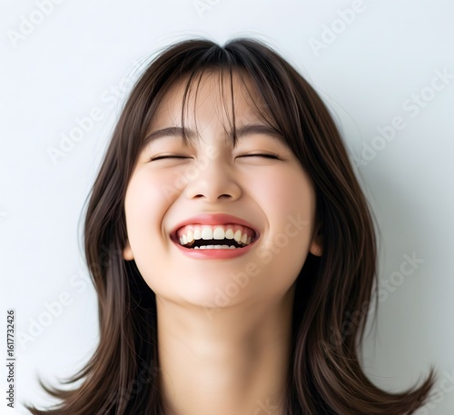 Joyful young East Asian woman with a radiant smile laughing heartily with her eyes closed on a clean background