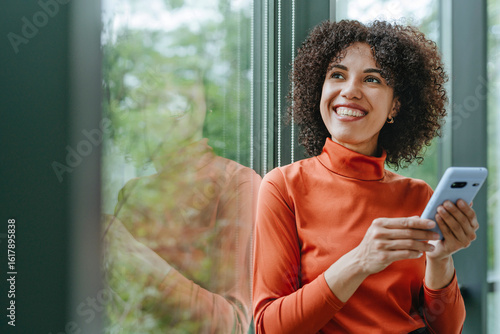 Businesswoman in orange turtleneck using phone and smiling by window