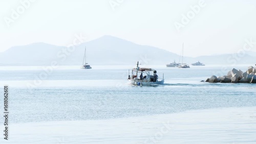 Wallpaper Mural Calm sea morning in Varkiza, Greece. A small fishing boat with people moves away near rocky breakwater. Moored dark yacht and distant hills visible through soft haze on horizon. Torontodigital.ca
