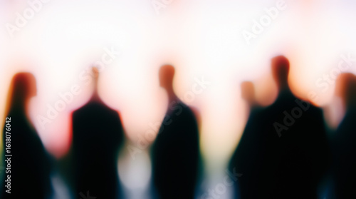 Blurred human figures stand against a bright gradient of pink, orange, and white, forming a mysterious abstract scene