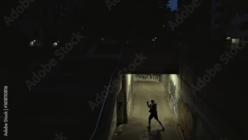 A lone figure in a hoodie and shorts practices boxing in a dimly lit, graffiti-covered underpass, embodying resilience and determination amidst urban grit, captured with a wide shot.
