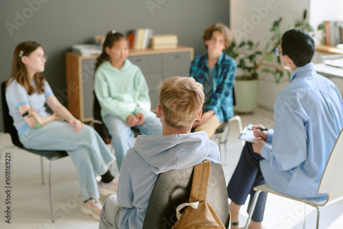 Group of multiethnic teenagers sitting in circle participating in discussion with Caucasian male counselor holding clipboard during therapy or support session indoors