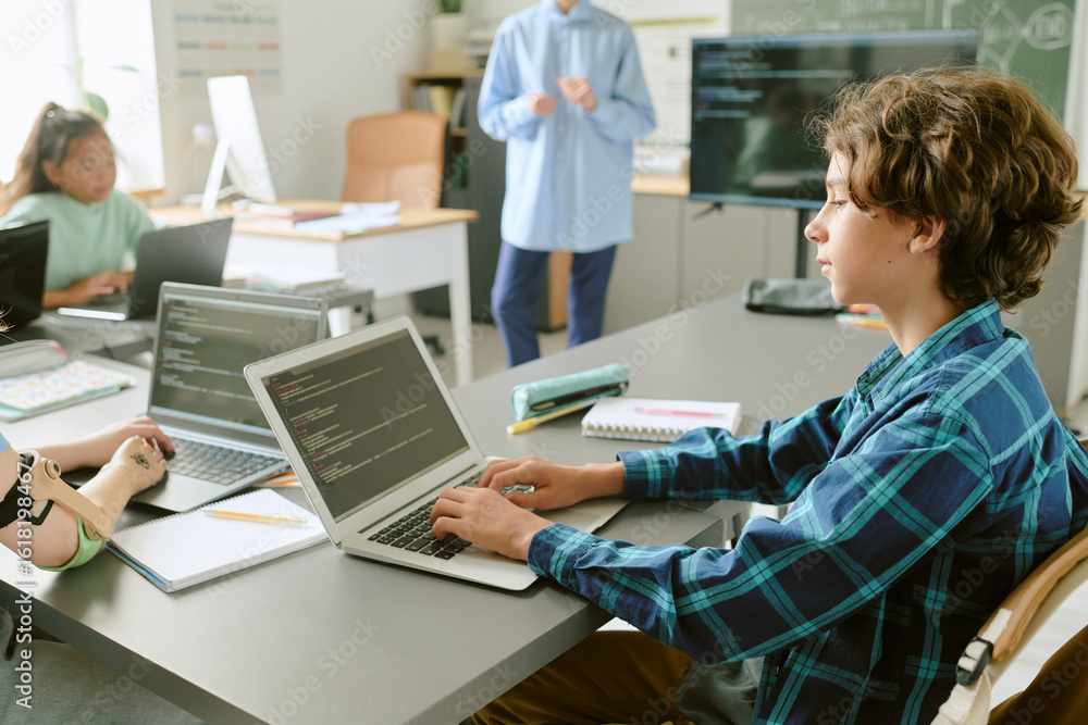 Obraz premium Teenage Caucasian boy working on laptop with code on screen in classroom, surrounded by classmates including Asian girl and student with prosthetic arm, teacher standing near chalkboard