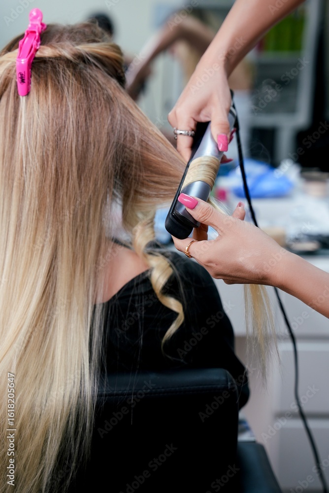 Fototapeta premium Hair stylist curling blonde hair with a flat iron in a beauty salon. Back view of a woman with half-straight and half-curled hair during hair styling process. Hands with hair tool visible