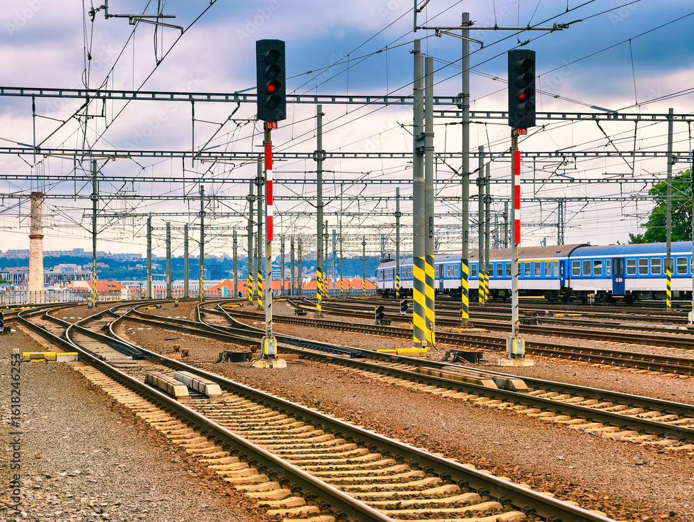 Fototapeta premium Railway Tracks and Overhead Power Lines at Train Station – Abstract Industrial Background with Red Signals and Blue Passenger Train, Ideal for Wallpaper, Web Design, Advertising, and Digital Use