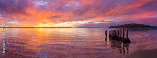 Panorama view of a colourful sunrise reflected in a calm ocean bay with wooden groynes