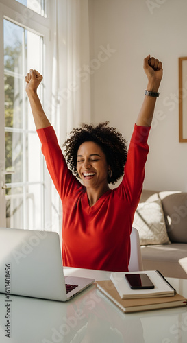 Ecstatic young woman celebrates professional success while working remotely from her bright home office, achieving goals and feeling a sense of accomplishment