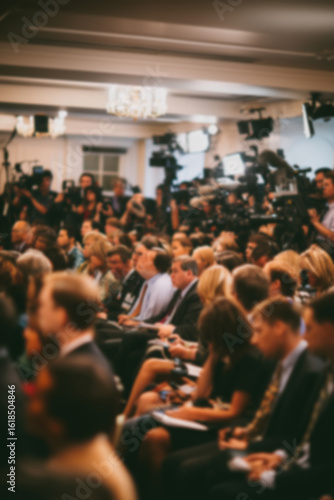 Blurred background image of a crowded press conference room filled with journalists seated and standing, with notepads, cameras, and microphones, all focused on a speaker. News event concept.