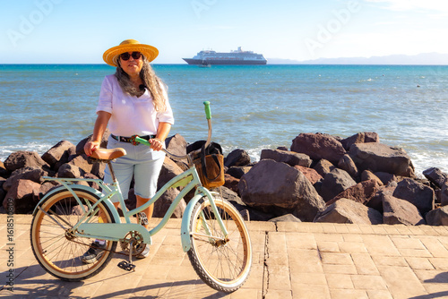 Female tourist standing next to bicycle on boardwalk, sea and cruise ship in background, wearing shorts, white blouse, hat and sunglasses, sunny day in Loreto, Baja California Sur, Mexico