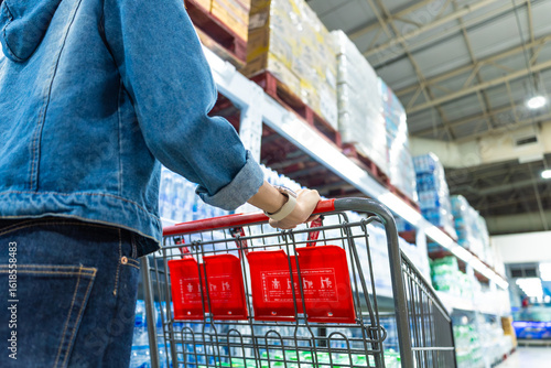 Image of asian woman shopper pushing a red shopping cart in a warehouse store aisle, surrounded by shelves full of bottled water and other items ready for customers to add to their carts.