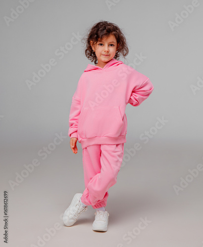 A child stands with one hand on her hip, wearing a vibrant pink tracksuit and white sneakers. She displays a playful expression against a simple backdrop.