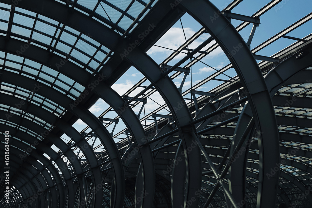 Fototapeta premium Modern steel roof structure closeup. Curved metal beams and glass panels of industrial building construction with sky background. Contemporary architectural detail of large station or commercial hall.