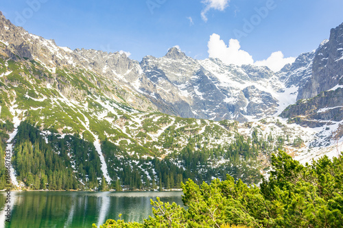 alpine lake in spring on a sunny day. travel landscape of rysy ridge in high tatra mountains of poland. snow among rocks and spruce forest on the hillside reflection in water. blue sky with clouds