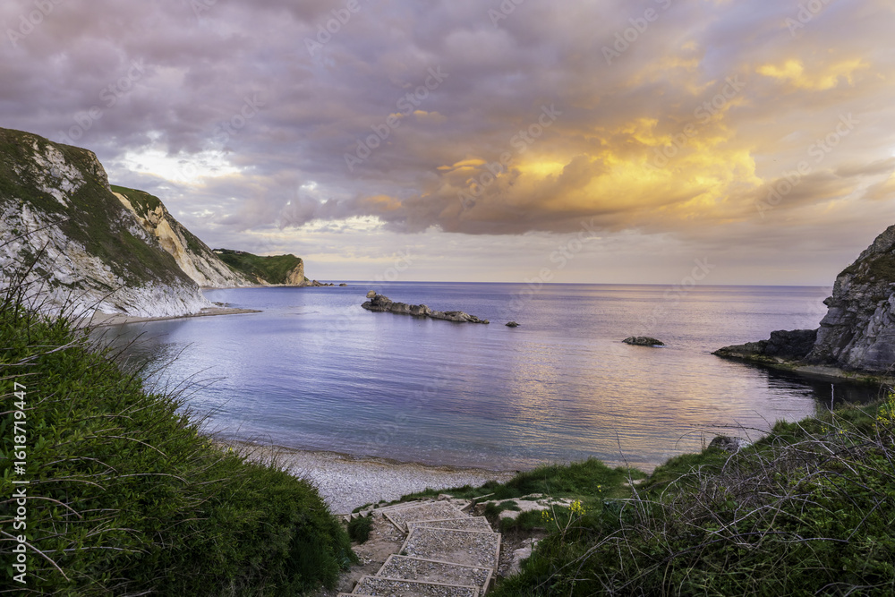View of dramatic cliffs embrace a tranquil bay as the sun paints the sky with vibrant hues of orange and purple, Wareham, England, United Kingdom. View of dramatic cliffs embrace a tranquil bay as the sun paints the sky with vibrant hues of orange and purple, Wareham, England, United Kingdom.