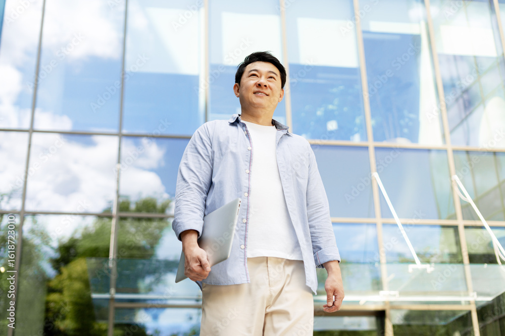 Asian businessman holding laptop looking up in front of modern office building Asian businessman holding laptop looking up in front of modern office building