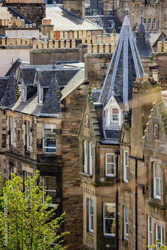 Unique and historic rooftops of Edinburgh's Old Town, Scotland, under a bright, partly cloudy sky. The foreground is dominated by the traditional stone buildings with their distinctive chimneys