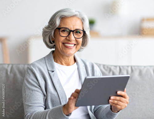 Smiling elderly woman with gray hair and glasses sitting on a modern sofa and using a digital tablet. Bright natural lighting, cozy home interior, photo-realistic scene.