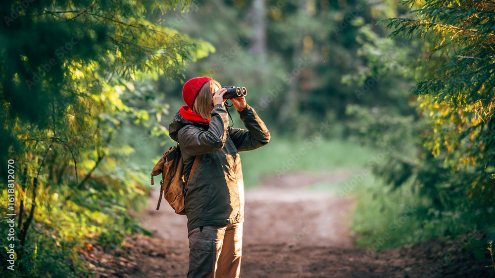 Fototapeta premium A middle-aged girl in a red hat and a green jacket stands in the forest on a sunny day and looks through binoculars into the distance,banner