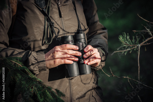 Fotografie A girl in green jacket stands in the forest on a sunny day and holds binoculars in his hands