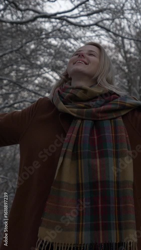 Vertical video of blonde European girl in coat and scarf rejoicing in snow within winter forest, capturing her delight
