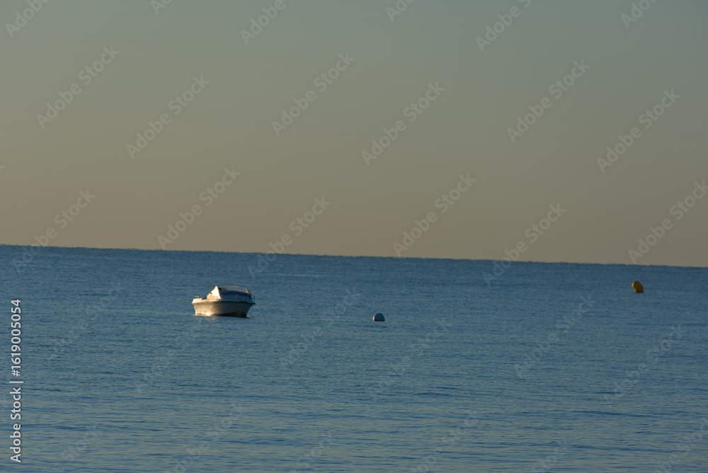 Fototapeta premium A small white motorboat with a light-colored canvas cover floats serenely on the calm blue water under a pale, muted sky.