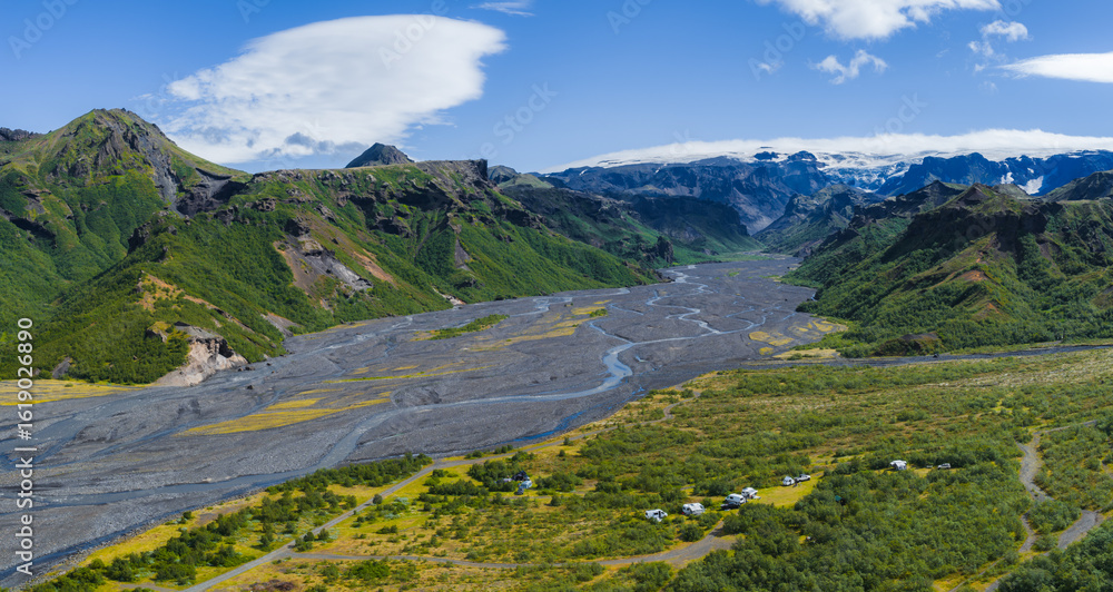 Obraz premium Aerial view of a green valley in Iceland with a braided river, rugged mountains, a glacier, and a campsite with vehicles and tents in the foreground.