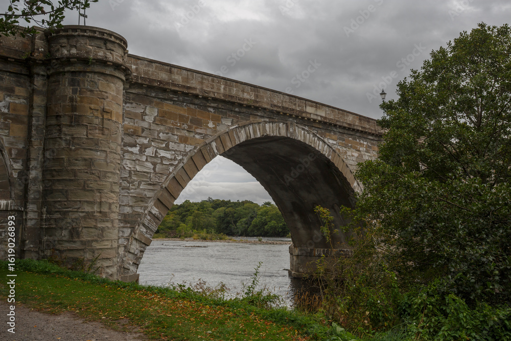 Historic stone bridge arching over a serene river under a cloudy sky in a natural landscape during autumn season Historic stone bridge arching over a serene river under a cloudy sky in a natural landscape during autumn season