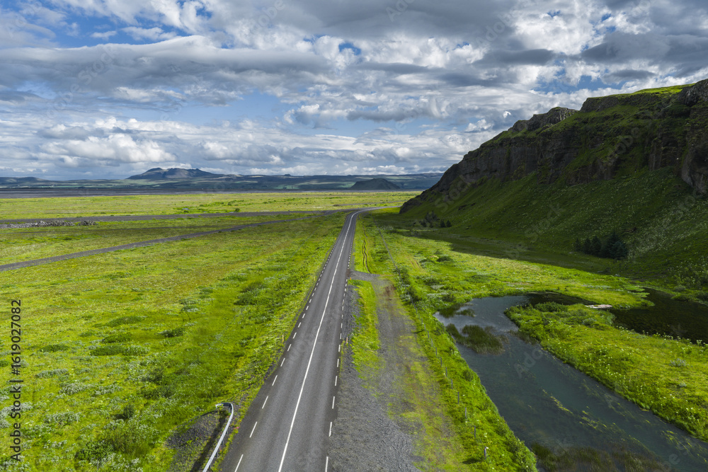 Naklejka premium A paved road extends through a green landscape in Iceland, bordered by a stream and grassy hillside, with mountains and clouds in the distance.