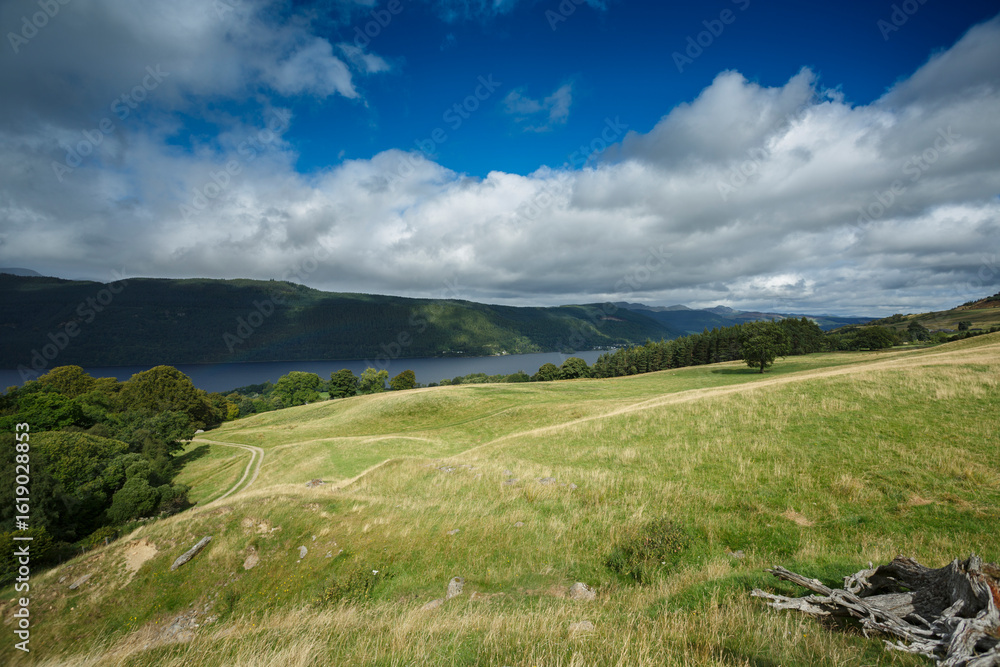 Fototapeta premium Expansive green landscape overlooking a tranquil lake surrounded by lush hills under a dynamic cloud-filled sky