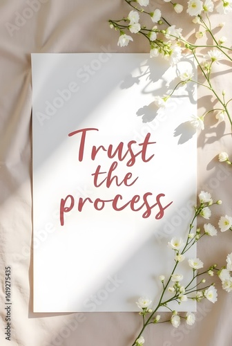A calming image featuring a white card with the inspirational phrase Trust the process written in a soft red hue, vertical composition. Delicate white flowers and soft sunlight create a peaceful.