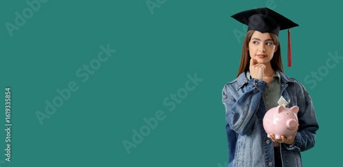 Thoughtful female student in graduation hat holding piggy bank on green backg...